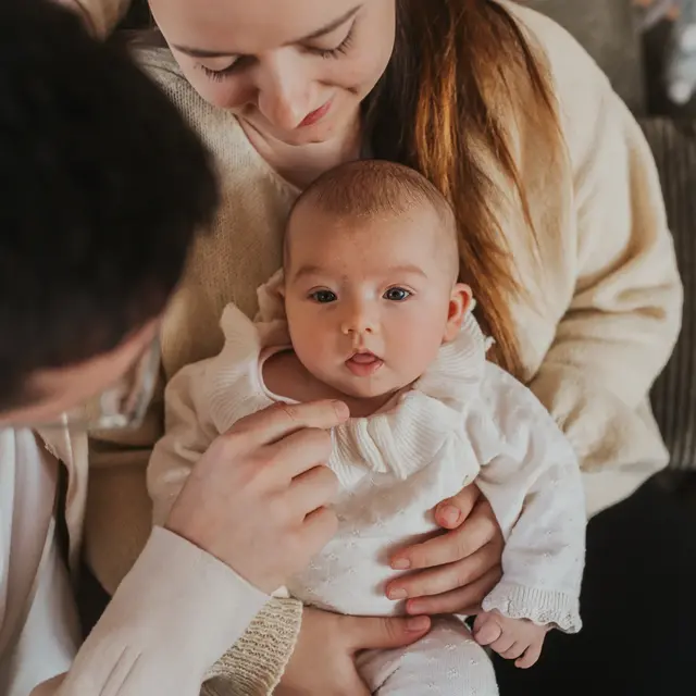 Mama und Papa betrachten ihr Baby im weißen Rüschenstrampler, das neugierig in die Kamera schaut