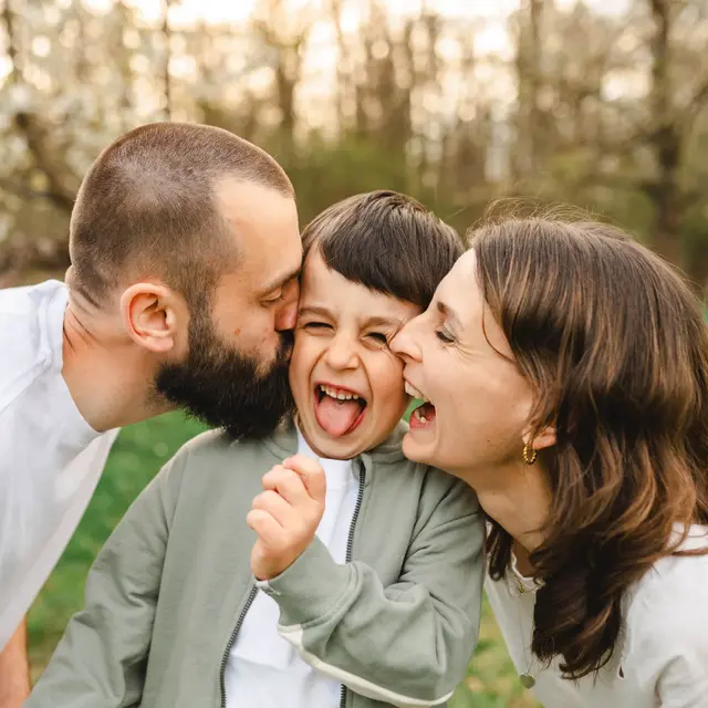 Mama und Papa küssen ihren lachenden Sohn gleichzeitig auf beide Wangen, dahinter blühende Bäume